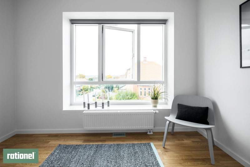 Lyst rum med trægulv, stol og grøn plante ved et stort energivenligt Rationel vindue, der giver udsigt til et hus med tegltag – et eksempel på løsninger fra energitjek